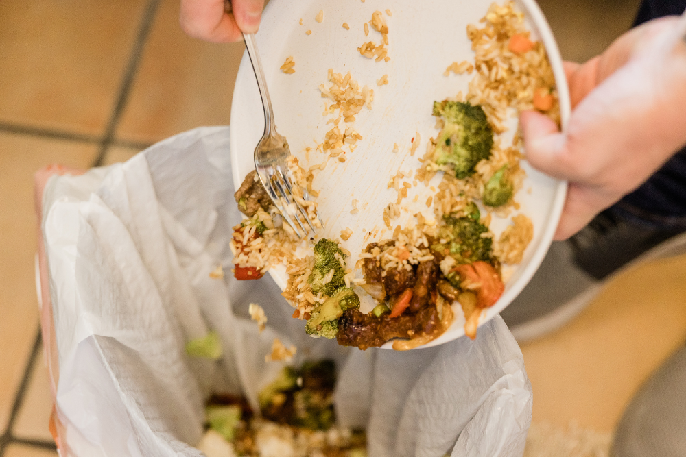 A close-up of a person scraping food off a plate into a garbage can.