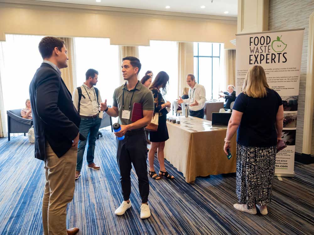 Two men are speaking to one another in a hotel hallway with an exhibit table and additional people behind them.