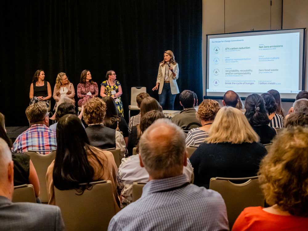 A woman is speaking to an audience with a microphone, while four other female panelists sit in chairs on a small stage.