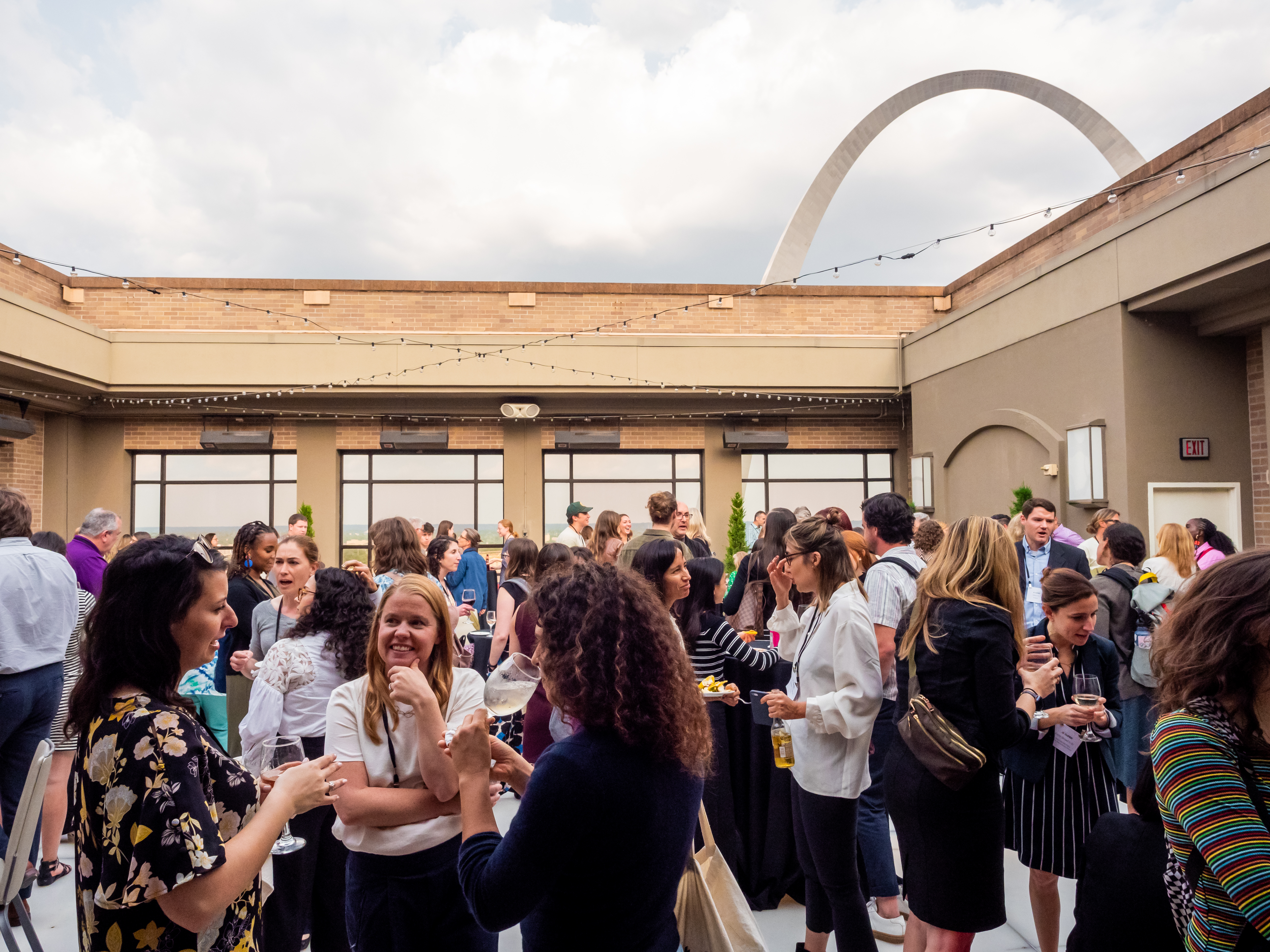 A group of people are standing and talking at a reception in an outdoor hotel space with the St. Louis Gateway Arch in the background.