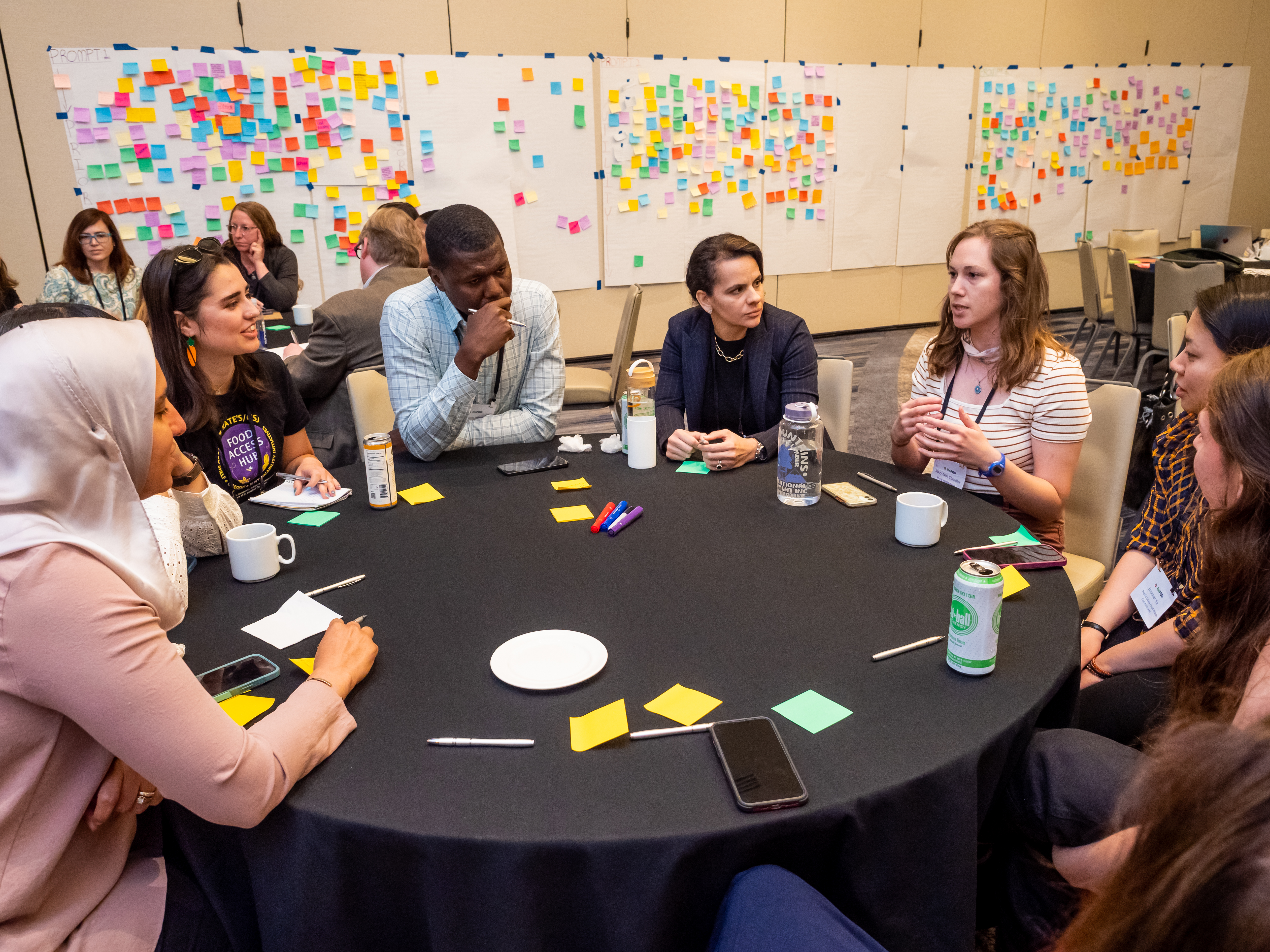 A group of people are sitting at a table and talking about food waste in a room featuring white sheets of paper covered with colorful sticky notes.